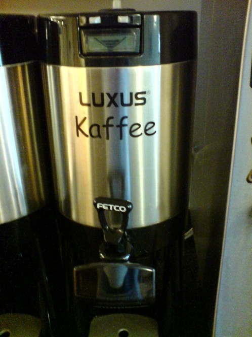 Eine Gastro-Thermoskanne in einem Frühstücksraum. Auf dem Edelstahlzylinder steht direkt unter dem Markennamen "Luxus" das Wort "Kaffee".